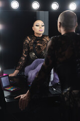 man in black lace dress and spectacular makeup looking in mirror in dressing room.