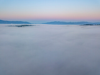 Naklejka na ściany i meble Flight over fog in Ukrainian Carpathians in summer. Mountains on the horizon. A thick layer of fog covers the mountains with a continuous carpet. Aerial drone view.