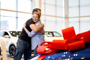 Male Shows New Car To Girlfriend. Present Concept. Hugging. Automobile Salon. Make A Decision. Gift Ribbon. Good Offer. Happy Together. Successful Buying. man makes a gift car to wife. in good mood.
