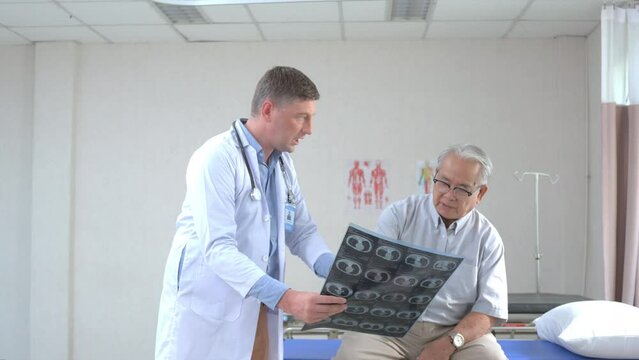 Selective focus of Caucasian male doctor in a white gown, pointing and explaining the abdomen MRI scan result to Asian senior male patient sitting on an examination bed in a hospital examination room.