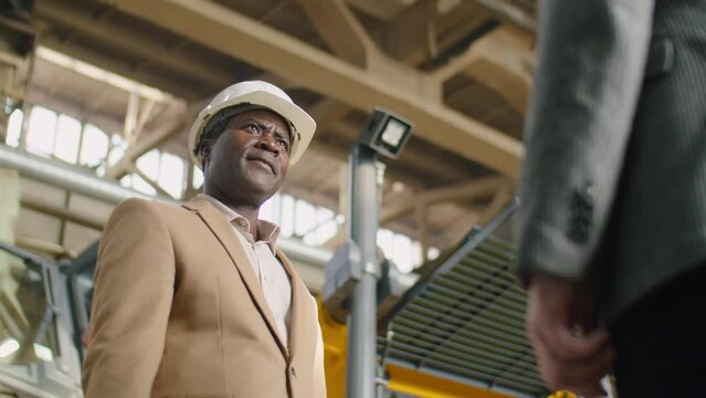 Low angle shot of middle aged African American engineer in hardhat and formalwear giving handshake to colleague in factory