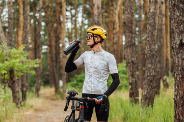Positive male cyclist standing in the woods with a bicycle dressed in sports equipment and drinking water from a bottle with a smile on his face. - Powered by Adobe