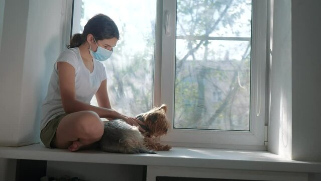 coronavirus pandemic concept. little teenage girl sitting in a medical mask at the window with a shaggy dog. self-isolation concept virus covid 19 infection doomsday. Sick child kid indoors. lifestyle