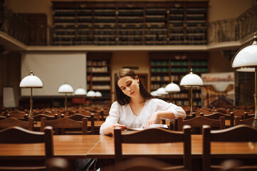 Beautiful girl in a white shirt sits at a table in a cozy old library and studies, reads books with a serious face.