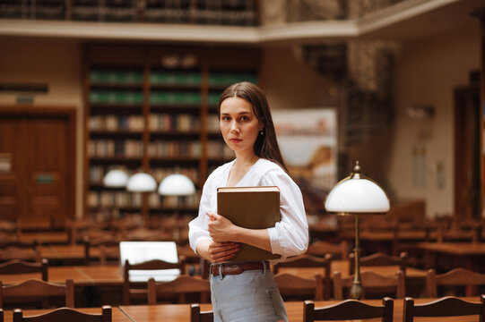 Attractive woman in a white blouse with a book in her hands stands in the library and looks at the camera with a serious face.