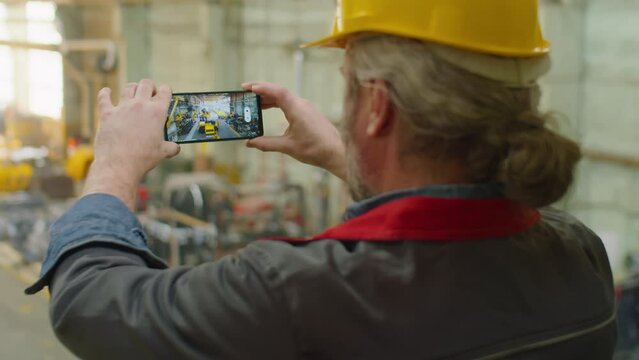 Factory quality control inspector in hardhat and workwear taking picture of machinery with smartphone