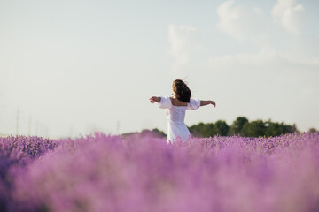 Young happy woman in white dress with hands up looking on lavender field and enjoying beautiful...
