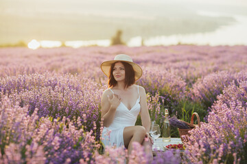 Elegant woman in white dress and straw hat on the lavender field having picnic at sunset. A girl on a picnic in the flowering lavender. River on the background.