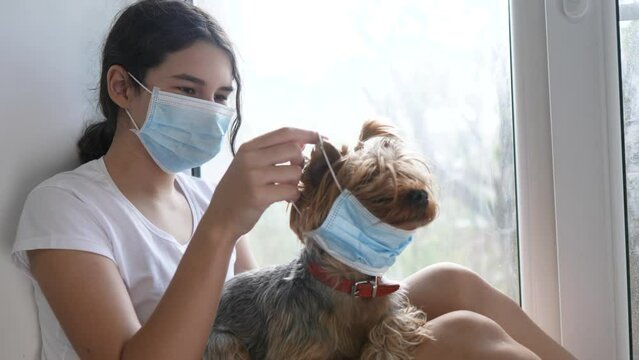 coronavirus pandemic concept. little teenage girl sitting in a medical mask at the window puts on a mask to the dog. self-isolation concept virus covid 19 infection doomsday. sick child kid indoors