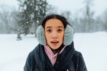 Portrait of surprised hispanic woman in winter clothes outdoors on a snowy day, looking shocked at the camera with open mouth on a background of winter views in the park.