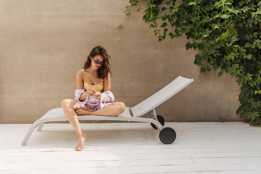 Portrait of cute young caucasian woman sitting on sun lounger in sunglasses against grey wall. Brunette with loose hair is resting on weekend. Summer lifestyle concept.