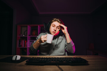 Tired sleepy gamer in a headset on his head with a cup in his hands looks at the computer screen playing online games at night at home. © bodnarphoto