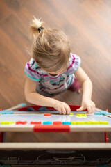  a 2-year-old child stands on a brown parquet and lays out magnetic numbers on the board with interest. View from above