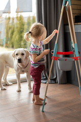 a little fair-haired girl stands near an easel by the window indoors, and a pet labrador is nearby