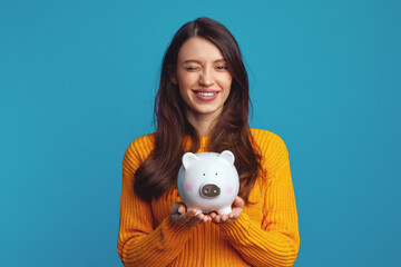 Cheerful young woman in casual orange sweater winking while holding white piggy bank with lots of...