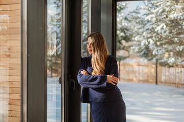stylish woman in a private house (barnhouse) wrapped herself in a warm knitted suit. Behind panoramic windows winter. Beautiful snowy winter outside the window.