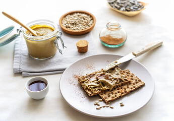 Homemade sunflower seed spread in jar with roasted seeds, maple syrup, cinnamon, crisp bread on light background
