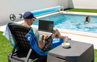Relaxed mature man working on a laptop and having an aperitif near the pool while his daughter is swimming
