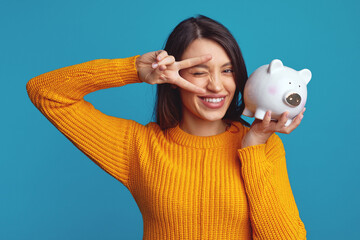 Excited young girl in casual orange sweater holding white piggy bank with lots of money, winking...