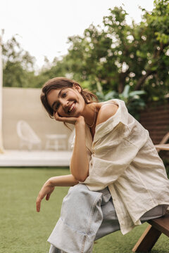 Beautiful young caucasian woman smiling looking into camera, sitting on bench, resting head on hand. Girl with gathered dark hair in shirt and jeans. Beauty, human emotions and spring holiday concept.