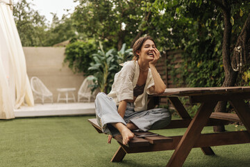 Cheerful young caucasian brunette lady is sitting on bench at table in backyard. Pretty woman in casual spring clothes spends her leisure time. Entertainment and lifestyle concept - Powered by Adobe