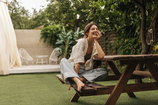 Cheerful young caucasian brunette lady is sitting on bench at table in backyard. Pretty woman in casual spring clothes spends her leisure time. Entertainment and lifestyle concept