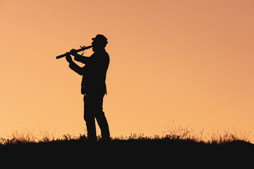 Fototapeta na wymiar silhouette of a man in a hat playing the flute. musician in nature outdoors against the backdrop of sunset
