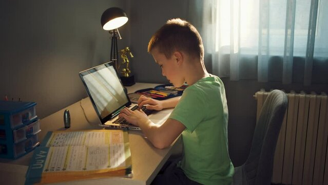 Boy sitting at the desk doing homework at home using laptop.