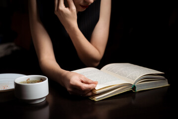 A woman is reading a book in the dark. Sits by the table.