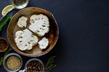 Cauliflower steak cooking. Raw cut cauliflower lie in a frying pan. Olive oil, herbs, various spices nearby. Dark background. Place for text.