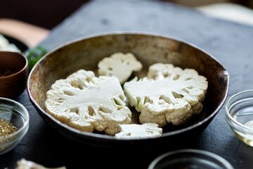Cauliflower steak cooking. Raw cut cauliflower lie in a frying pan. Olive oil, herbs, various spices nearby. Dark background. Place for text.