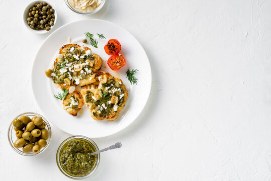 Cauliflower steak with spices, chimichurri sauce, almond flakes, olives, fried cherry tomatoes and capers on a white plate. Vegetarian food. White background. Place for text.