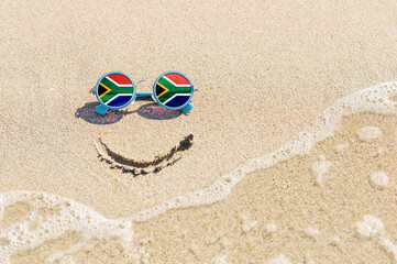 Fototapeta na wymiar A painted smile on the sand and sunglasses with the flag of South Africa. The concept of a positive and successful holiday in the resort of South Africa.