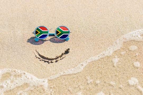 A Painted Smile On The Sand And Sunglasses With The Flag Of South Africa. The Concept Of A Positive And Successful Holiday In The Resort Of South Africa.