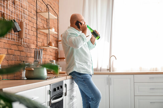 Bald man drinking beer and talking by phone in kitchen