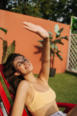 Smiling caucasian young woman hiding from rays, light from sun on face and shadows from hands. She is wearing summer top and her hair is loose. Relax, leisure at home, outdoors.