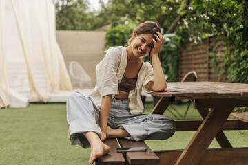 Funny fair-skinned young woman with beaming smile looks at camera while sitting on bench. Natural makeup, dark hair. She is wearing top, shirt and jeans. Concept of solitude with nature.