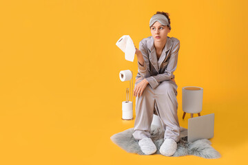 Young woman in pajamas and with paper sitting on toilet bowl against color background
