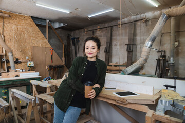 Cheerful carpenter holding coffee to go and looking at camera in workshop.