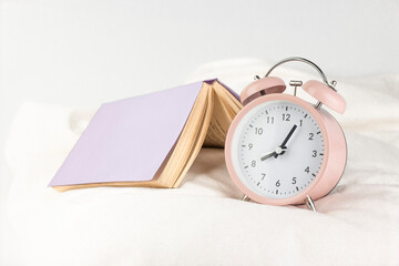 Pink alarm clock with opening book on the bed close up image. Morning concept.