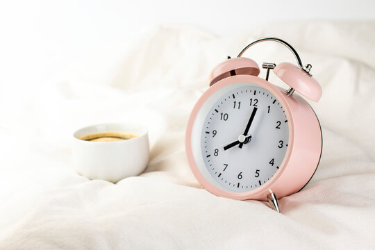 Morning concept. Pink alarm clock with cup of coffee on the bed close up image.