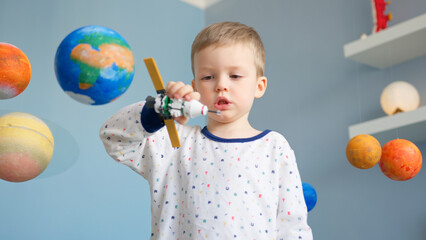 Portrait blond boy is playing with space satellite made by constructor, flying in space near...