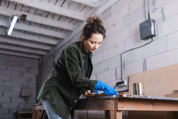 Carpenter in shirt and rubber glove applying wood stain on plank in workshop.