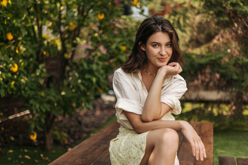 Young pretty fair-skinned lady is playfully looking at camera while sitting in orchard alone. Dark-haired model wears light-colored casual summer clothes. Lifestyle, emotions, leisure concept.