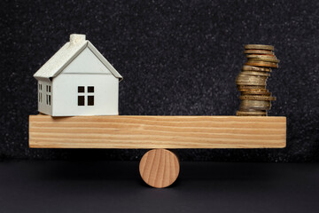 white metal house and a stack of coins balanced on a seesaw, on dark background