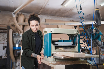 Brunette woodworker standing near thickness planer in workshop. - obrazy, fototapety, plakaty
