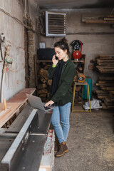 Brunette carpenter talking on smartphone and using laptop in workshop.
