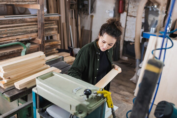 Fototapeta na wymiar Brunette carpenter holding wooden board near thickness planer and planks in workshop.