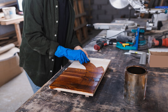Cropped view of furniture designer in rubber glove applying wood stain on board in workshop.
