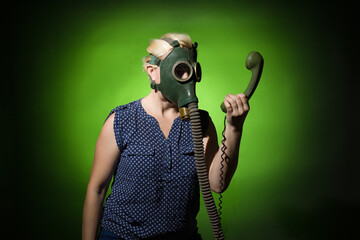 Funny woman in a gas mask looks in surprise at the handset of a retro phone on a dark background,...